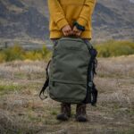 Photo d'une personne en veste jaune et sac à dos noir marchant dans un champ d'herbe brune en plein jour