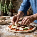 Chef préparant une pizza artisanale au jambon, fromage, mozzarella et champignons dans une cuisine rustique.